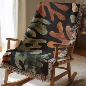 May include: A woven throw blanket draped over a wooden rocking chair. The blanket features a black background with abstract shapes in shades of green, brown, and orange. The edges are finished with a fringe.