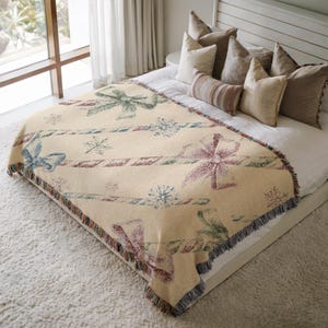 May include: A cream-colored throw blanket with a repeating pattern of bows, candy canes, and snowflakes. The bows are in shades of blue, green, and burgundy. The blanket has a fringed edge and is displayed on a bed.