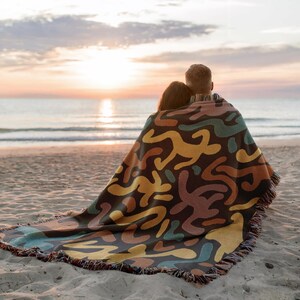 Pu&ograve; includere: Una coperta fantasia con bordo a frange copre due persone su una spiaggia sabbiosa. La coperta presenta un motivo astratto ripetuto nei toni del marrone, giallo, turchese e arancione. Il mare e il tramonto sono visibili sullo sfondo.