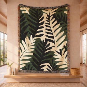 May include: A woven throw blanket featuring a repeating pattern of large fern leaves in shades of dark green, black, and cream. The rectangular textile has a textured surface and is finished with a decorative fringed edge on all four sides.