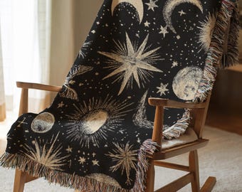 May include: A black throw blanket with a celestial design featuring moons, stars, and suns in a cream color. The blanket is draped over a wooden rocking chair and has a fringed edge.
