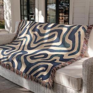 May include: A woven throw blanket with a cream-colored background and a navy blue, abstract, swirled pattern. The blanket is draped over a white wicker sofa. The edges are finished with a fringe in shades of red, orange, and brown.