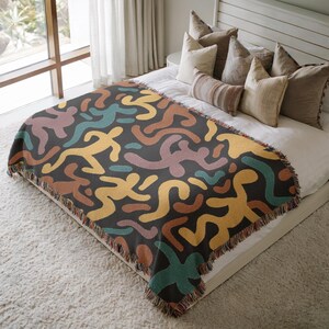 Pu&ograve; includere: Una coperta grigio scuro con un motivo ripetuto di figure umane astratte in giallo, verde acqua, marrone e viola. La coperta ha un bordo con frange ed &egrave; esposta su un letto con cuscini.