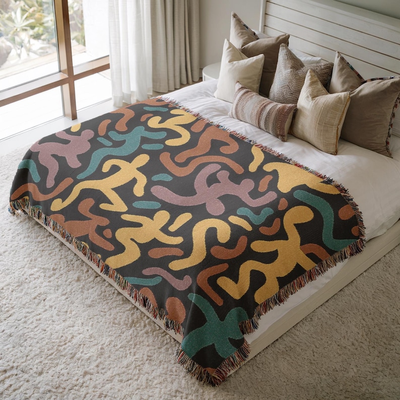 Pu&ograve; includere: Una coperta grigio scuro con un motivo ripetuto di figure umane astratte in giallo, verde acqua, marrone e viola. La coperta ha un bordo con frange ed &egrave; esposta su un letto con cuscini.