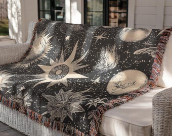 May include: A dark gray throw blanket with a celestial design featuring suns, stars, and planets in cream and white. The blanket has a fringed edge and is draped over a white wicker sofa.