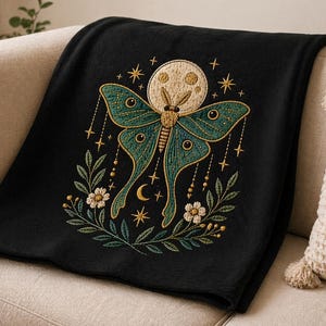 May include: Black throw blanket featuring an embroidered design of a luna moth with teal wings, a cream-colored moon, and gold stars. The moth is surrounded by floral and leaf accents. The blanket is draped over a beige sofa.