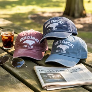 May include: Three baseball caps in shades of burgundy, navy, and light blue, each bearing the text "NORTH CAROLINA EST. 1789" and a white outline of the state. The caps are stacked on a wooden picnic table, alongside sunglasses and a newspaper.