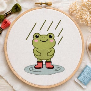 May include: An embroidery hoop features a cartoon frog in the rain, wearing red boots and standing in a puddle. The frog is green with pink cheeks and black eyes. Embroidery floss in green and coral, and a spool of blue thread are also visible.