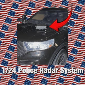 Op de afbeelding: Een zwart politiemodelauto op schaal 1:24 met een radarsysteem op de motorkap. Een rode pijl wijst naar het radarsysteem. De achtergrond is een patroon van Amerikaanse vlaggen.