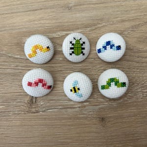 Könnte beinhalten: Sechs weiße, stoffbezogene Knöpfe mit gestickten Insekten- und Wurmdesigns. Die Designs umfassen einen gelb-orangen Wurm, einen grünen Käfer, blau-weiße Formen, einen roten Wurm, eine Biene und einen grünen Wurm.