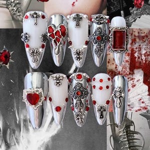 May include: A collection of ten long, metallic silver nail designs. Each nail features intricate embellishments, including crosses, hearts, and red gemstones. Some nails have a white base, while others are silver. The overall aesthetic is gothic and ornate.