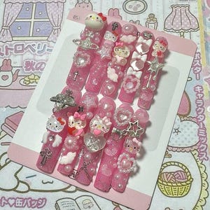 May include: A set of pink press-on nails with various embellishments. Each nail features glitter, pearls, and charms, including Hello Kitty figures, hearts, bows, and stars. The nails are arranged on a pink card.