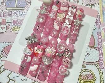 May include: A set of pink press-on nails with various embellishments. Each nail features glitter, pearls, and charms, including Hello Kitty figures, hearts, bows, and stars. The nails are arranged on a pink card.