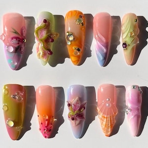 Puede incluir: Una colección de diez uñas artificiales largas y ovaladas. Cada uña presenta un diseño único, incluyendo acentos florales, gemas y patrones abstractos. Los colores varían desde rosas y naranjas hasta verdes y morados, con un acabado brillante.