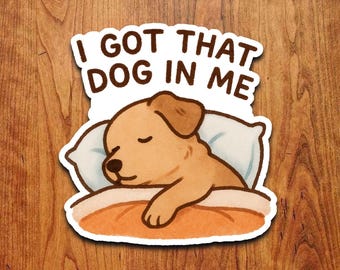 May include: A sticker featuring a sleeping cartoon dog with the text "I GOT THAT DOG IN ME". The dog is light brown and resting on a pillow and blanket. The sticker has a white border and is set against a wood grain background.