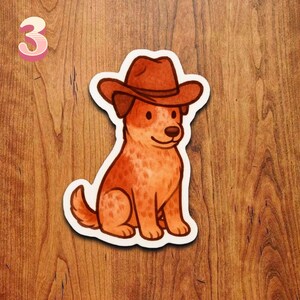 May include: A sticker featuring a cartoon dog wearing a brown cowboy hat. The dog is sitting with its tail curled and has a reddish-brown coat. The number "3" is in the upper left corner, in a pink and white font.