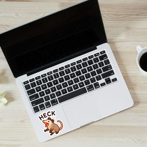 以下が含まれることがあります： 黒い画面とキーボードを備えたラップトップ、コーヒーカップ、鉛筆が明るい木製の表面に置かれています。漫画のオポッサムと「HECK」という文字が書かれたステッカーがラップトップに貼られています。