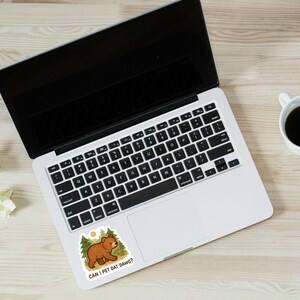 May include: Laptop with a sticker of a cartoon bear in a forest. The sticker reads "CAN I PET DAT DAWGG?". A coffee cup, pencils, and scrunched paper are on a wooden surface.