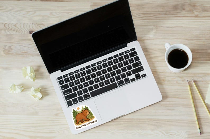 May include: Laptop with a sticker of a cartoon bear in a forest. The sticker reads "CAN I PET DAT DAWGG?". A coffee cup, pencils, and scrunched paper are on a wooden surface.