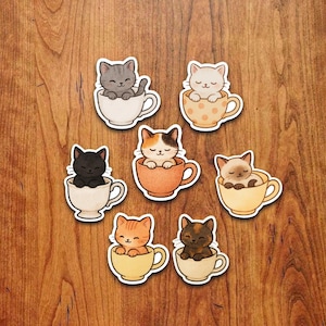 May include: A collection of seven cat stickers, each featuring a different colored cat nestled inside a teacup. The cats vary in color, including gray, black, orange, and calico. The teacups are in various colors, some with polka dots.