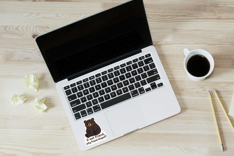 May include: Laptop with a black screen and keyboard, a sticker of a brown bear with the text "if not friend, why friend shaped?", a cup of coffee, pencils, and crumpled paper on a light wood surface.