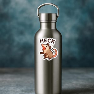 以下が含まれることがあります： 「HECK」の文字が上に書かれた、漫画のオポッサムのステッカーが付いたステンレス製のウォーターボトル。オポッサムは茶色で、ピンク色の尻尾と開いた口をしています。ボトルにはシルバーのハンドルが付いています。