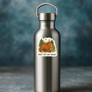 May include: A silver water bottle with a sticker depicting a brown bear in a forest setting. The sticker has the text "CAN I PET DAT DAWG?" in a fun font. The background is a blurred blue.