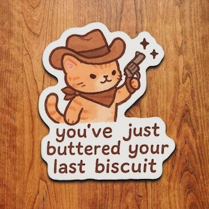 May include: A sticker featuring a cartoon orange tabby cat wearing a cowboy hat and bandana, holding a gun. The text "you've just buttered your last biscuit" is below the cat.