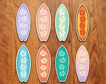 Könnte beinhalten: Acht Surfbrett-Aufkleber in verschiedenen Farben, darunter Lila, Rosa, Blau, Orange und Grün. Jedes Surfbrett hat ein Design mit weißen Hibiskusblüten. Die Aufkleber sind auf einer Holzoberfläche angeordnet.