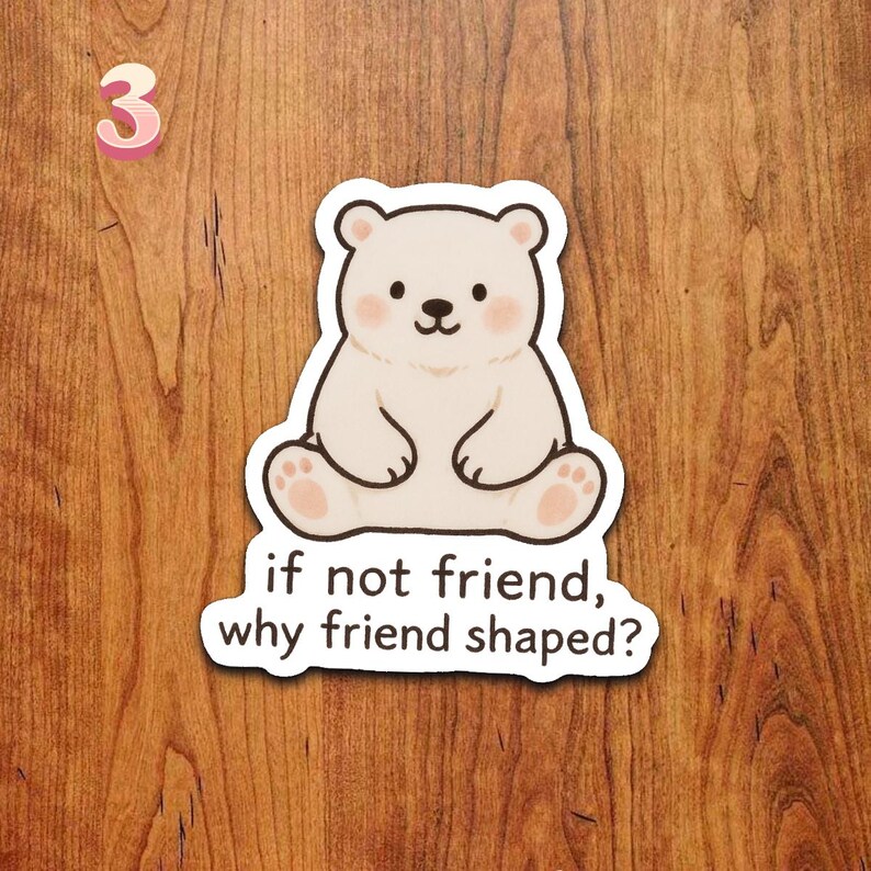 Puede incluir: Una pegatina blanca con un oso de dibujos animados con detalles rosas en las mejillas y las patas. La pegatina incluye el texto "if not friend, why friend shaped?" y el n&uacute;mero rosa "3". La pegatina est&aacute; sobre una superficie de madera.