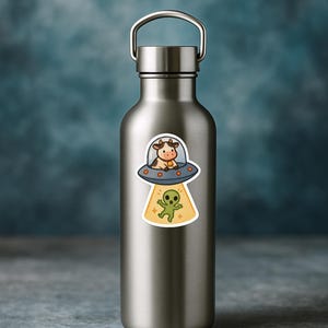 May include: A stainless steel water bottle with a sticker featuring a cartoon cow in a UFO above a green alien. The cow is brown and white, and the alien is green with a yellow glow.