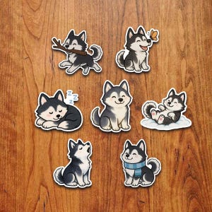 Op de afbeelding: Een verzameling van zeven cartoon husky stickers. Elke sticker toont een andere pose, waaronder een husky met een stok, een vlinder, slapend, zittend, rollend op een wolk, huilend en met een sjaal. De stickers zijn zwart, wit en grijs.