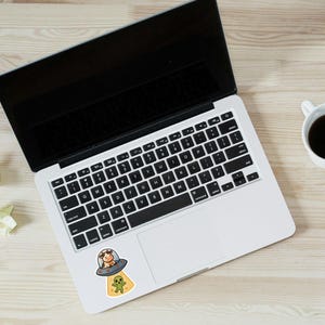May include: A laptop with a black screen and keyboard, a coffee cup, and pencils on a light wood surface. A sticker of a cow in a UFO with an alien is on the laptop.