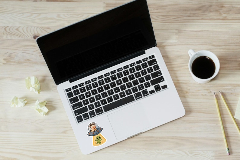 May include: A laptop with a black screen and keyboard, a coffee cup, and pencils on a light wood surface. A sticker of a cow in a UFO with an alien is on the laptop.
