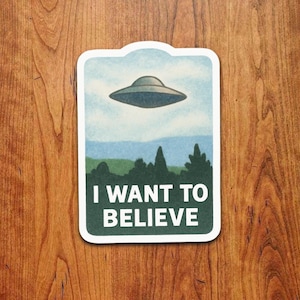 Può includere: Un adesivo rettangolare con bordo bianco presenta un'illustrazione retrò di un disco volante sopra una foresta e il testo "I WANT TO BELIEVE" in lettere bianche su sfondo verde. Lo sfondo è un cielo azzurro chiaro.