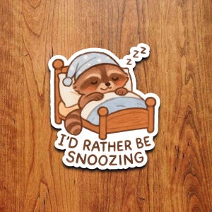 Può includere: Un adesivo con un procione dei cartoni animati che dorme in un letto, con un berretto da notte blu a pois bianchi. Il procione è marrone e beige con gli occhi chiusi. Il testo "I'D RATHER BE SNOOZING" è sotto il letto.