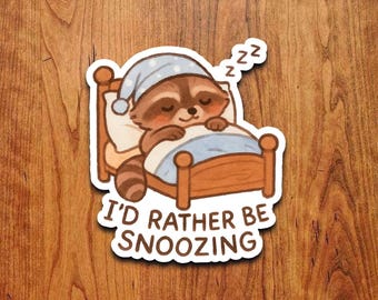 May include: A sticker featuring a cartoon raccoon sleeping in a bed, wearing a blue nightcap with white polka dots. The raccoon is brown and tan with closed eyes. The text "I'D RATHER BE SNOOZING" is below the bed.