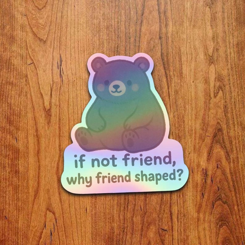 May include: A holographic sticker featuring a cartoon bear in rainbow colors. The bear is sitting down with a friendly expression. The sticker has the text "if not friend, why friend shaped?"