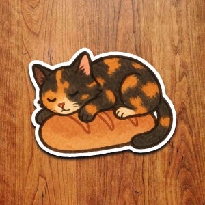 Op de afbeelding: Een sticker met een slapende lapjeskat met zwarte en oranje vlekken, liggend op een brood. De kat is opgerold, ogen gesloten, en het brood is goudbruin van kleur.