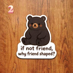 Puede incluir: Una pegatina con un oso negro de dibujos animados con una expresi&oacute;n amigable y mejillas rosadas. El oso est&aacute; sentado encima del texto "if not friend, why friend shaped?" El n&uacute;mero "2" est&aacute; en la esquina superior izquierda.