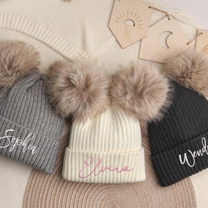 Puede incluir: Tres gorros de punto acanalado con grandes pompones de piel sintética. El gorro gris presenta el nombre Sophia bordado en blanco, el gorro crema tiene el nombre Elvira bordado en rosa y el gorro negro muestra el nombre Wendy bordado en blanco.
