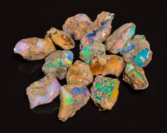 Puede incluir: Una colección de piedras preciosas de ópalo en bruto con colores vibrantes e iridiscentes. Los ópalos muestran destellos de azul, verde, naranja y rosa dentro de una matriz de roca marrón y marrón. Las piedras preciosas están dispuestas sobre una superficie negra.
