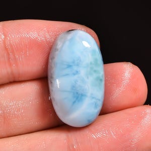 May include: Oval-shaped, polished gemstone with a light blue and white pattern, reminiscent of ocean waves. The stone has a glossy finish and is held in a hand, showcasing its natural beauty. The gemstone is a Larimar.