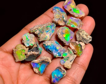 Könnte beinhalten: Eine Sammlung roher Opal-Edelsteine, die lebendige Farben wie Blau, Grün und Lila zeigen. Die Opale haben eine raue, natürliche Textur mit Erdtönen. Die Edelsteine werden in einer Hand vor schwarzem Hintergrund gehalten.