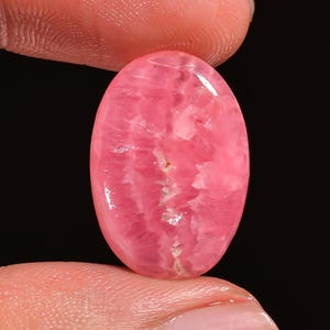 Op de afbeelding: Een ovaalvormige rhodochrosiet edelsteen met een gepolijst, glad oppervlak. De steen vertoont een levendige roze kleur met ingewikkelde, lichtere roze en witte kristallijne patronen die door de gehele structuur van de steen lopen.