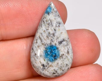May include: A teardrop-shaped K2 stone, held between fingers. The stone features a white and gray speckled pattern with a vibrant blue center and tip. The polished gemstone is a natural mineral.