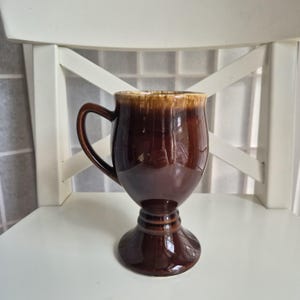 Pode incluir: Uma caneca de cerâmica marrom com base de pedestal e alça curva. A caneca tem um acabamento brilhante com uma borda marrom mais clara. A caneca está em uma cadeira branca.