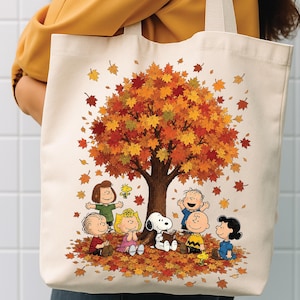 May include: A cream-colored canvas tote bag featuring a colorful graphic of the Peanuts characters gathered under a large autumn tree with falling orange, red, and yellow leaves. Snoopy is reading a book while surrounded by his friends.
