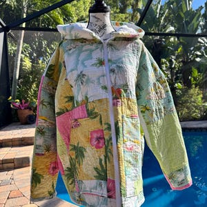 May include: A quilted zip-up hooded jacket featuring a patchwork design with tropical motifs, including palm trees, hibiscus flowers, and the text Aloha Hawaii. The garment has a white front zipper, long sleeves, and a light-colored interior lining.