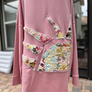 May include: A dusty pink long-sleeve sweatshirt featuring several asymmetrical, quilted fabric patches sewn onto the back. The patches display various floral and geometric patterns in shades of cream, green, yellow, and red with visible stitching.